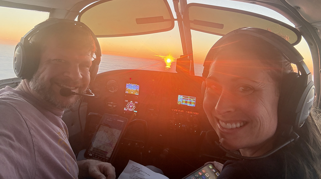 A man and a woman sit in the front seats of a small airplane with a colorful sunset visible through the windshield.