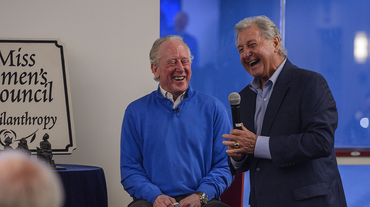 Two men, one holding a microphone, share a laugh on a stage.