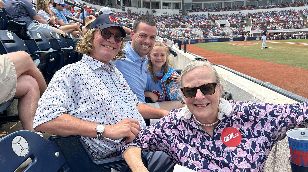 Two men, a woman and a girl sti in the stands at a baseball stadium.