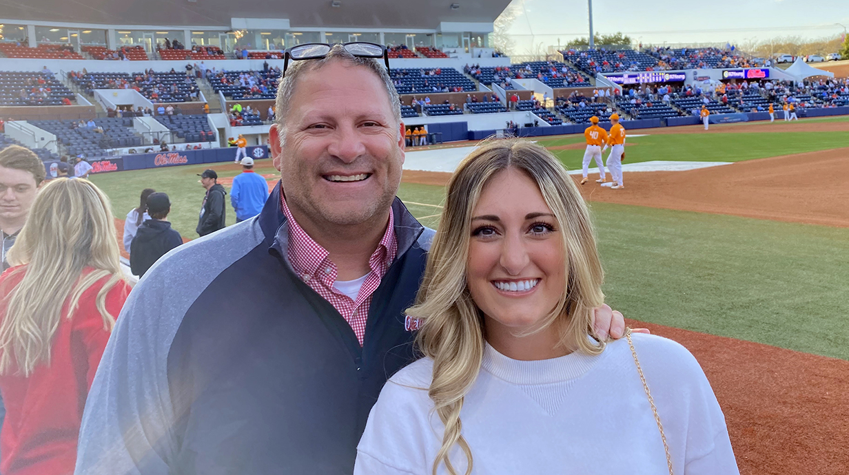 A man and a young woman stand in a baseball stadium with a game in progress behind them.