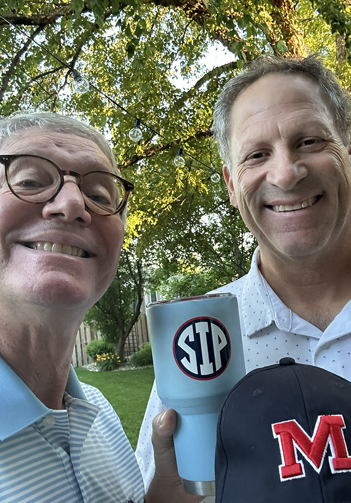 Two men stand outdoors, holding an Ole Miss baseball cap and travel mug.