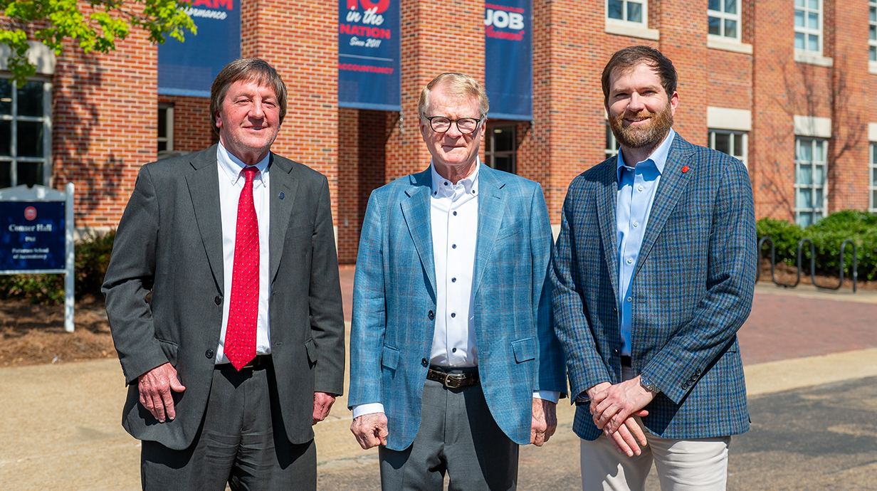 Three men wearing suits stand in the street outside a large brick building.