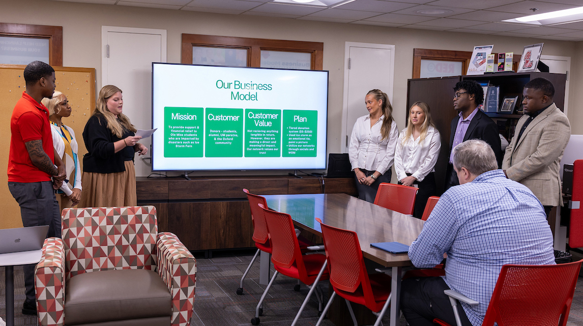A group of young people stand near a projection screen in the front of a meeting room while a man sitting at a table listens.