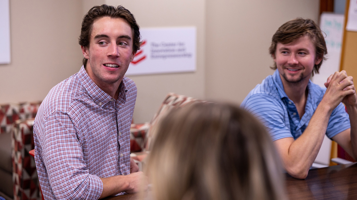 A young man seated at a meeting table while another young man listens.