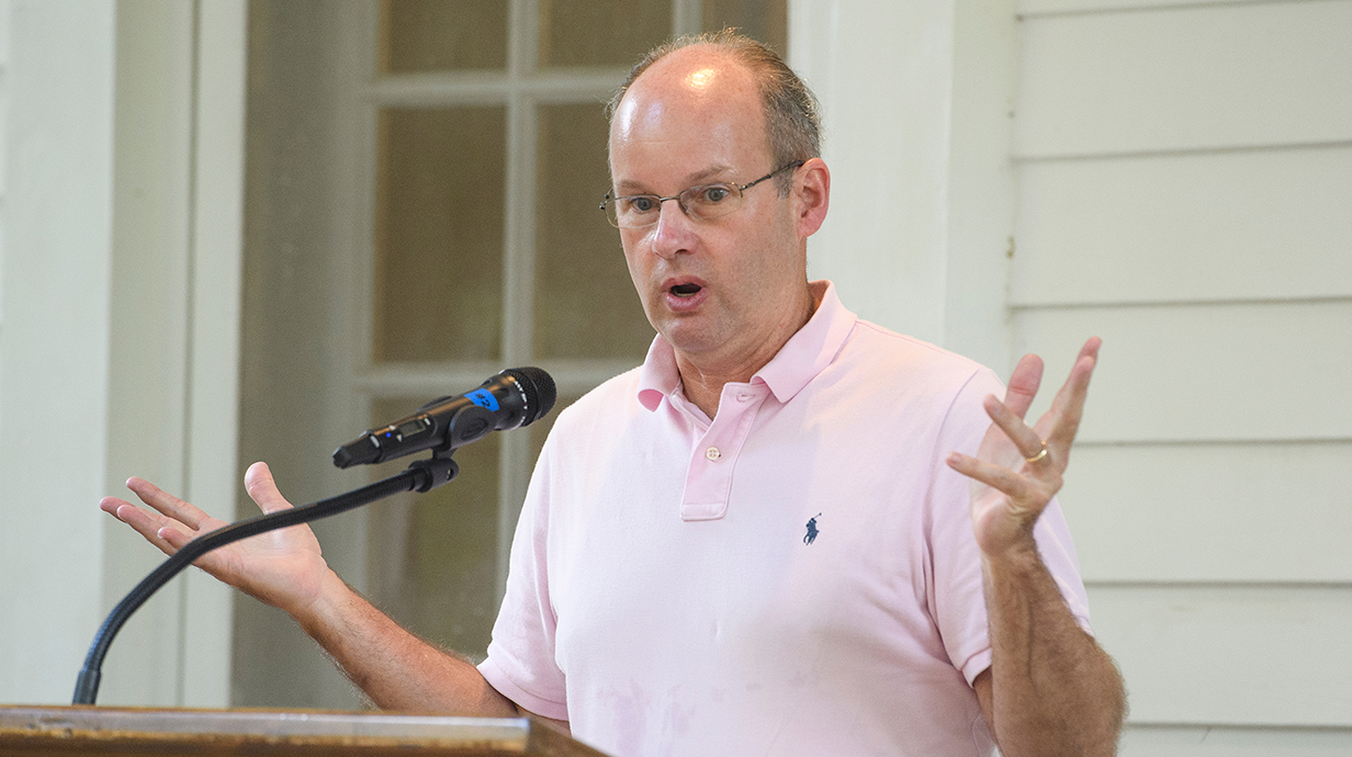 A man wearing a pink polo shirt gestures as he talks from an outdoor podium.