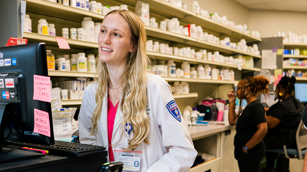 A young woman wearing a white pharmacists coat sits at a computer terminal while technicians work in the background.