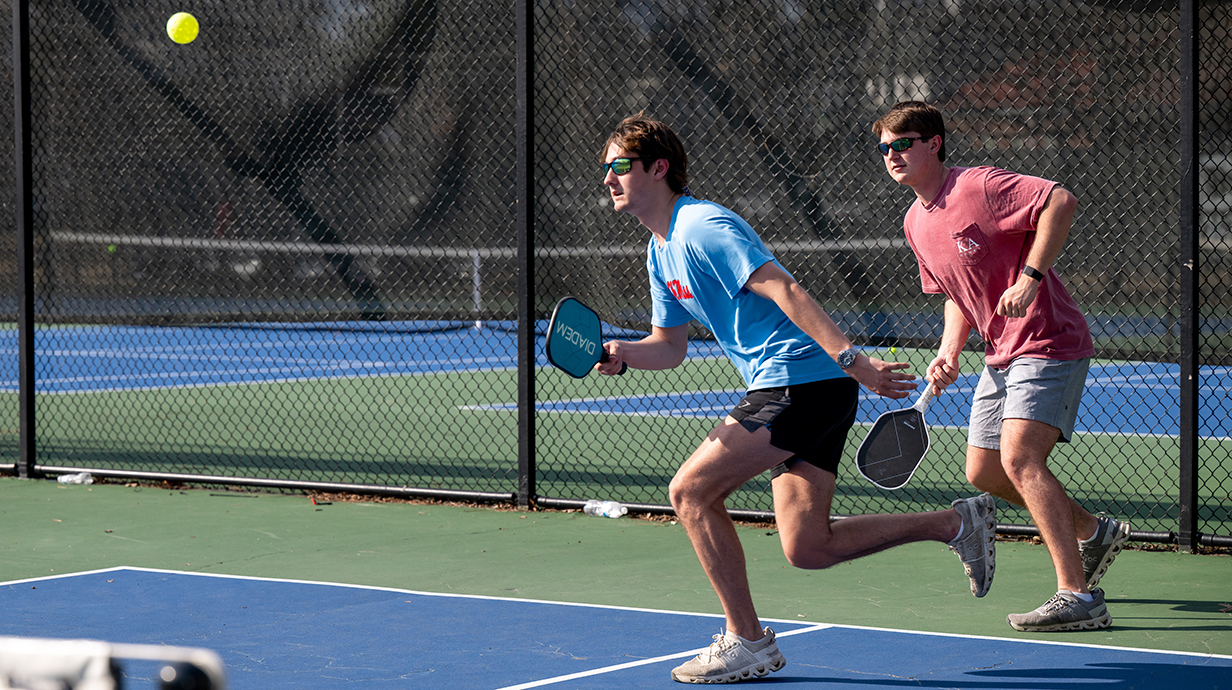 Two young men run across a pickleball court to return a shot.