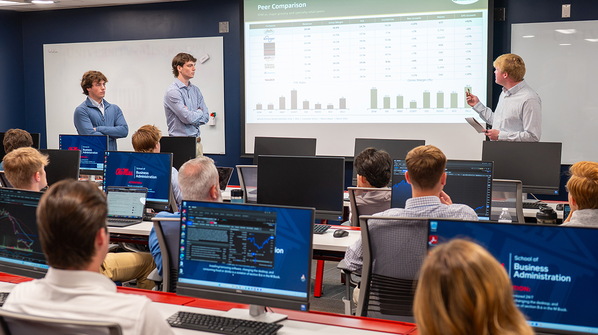 Three young men talk to a classroom of people from either side of a projection screen while other young people sit at computer terminals displaying financial information.