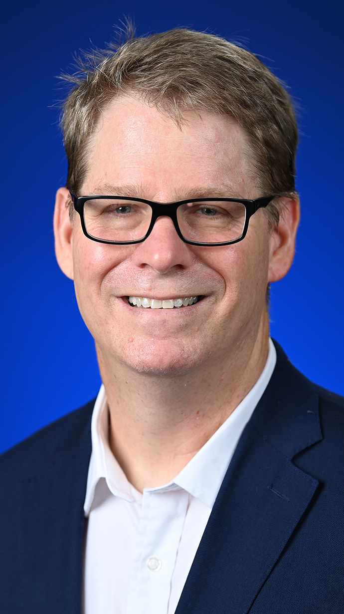 Headshot of a man wearing glasses and a navy blue jacket over a white shirt.