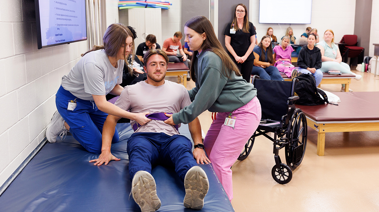 Two young women help a young man sit up from a padded bench in a physical therapy lab setting.