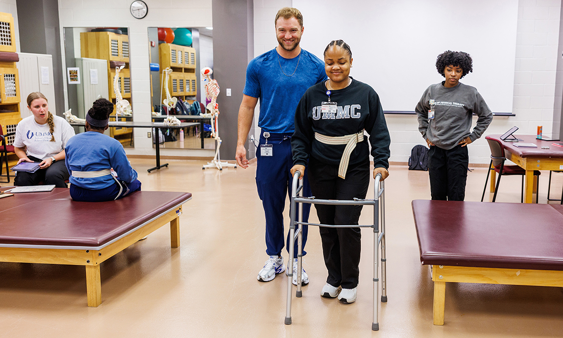 A young man help guide a young woman using a walker in a classroom.