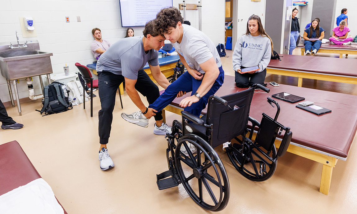 A young man helps another young man move from a bench to a wheelchair while a young woman watches nearby.