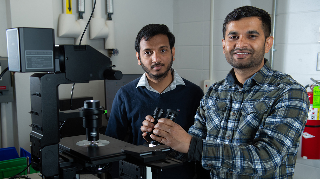 Two men stand in a laboratory, where one holds a microscope eyepiece.
