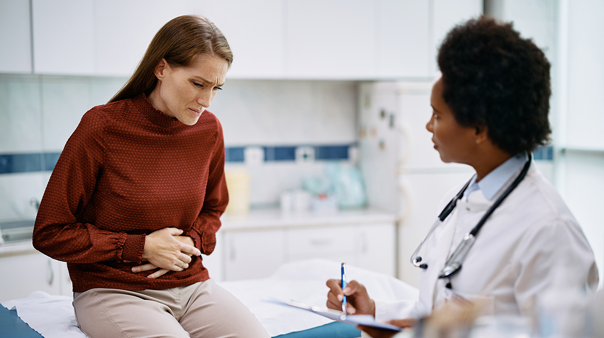 A woman sitting on an examining table holds her stomach in pain as a female doctor writes on a clipboard.