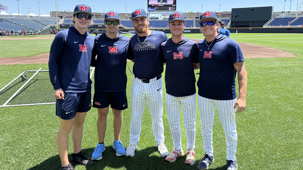 Five young men, all wearing navy blue Ole Miss shirts and baseball caps, stand on the infield of a baseball stadium.