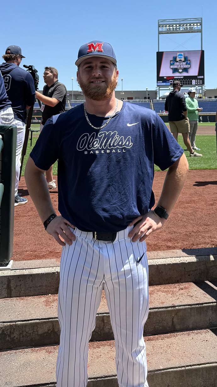 A young man wearing a navy blue Ole Miss shirt and baseball cap stands at the edge of the field in a baseball stadium.