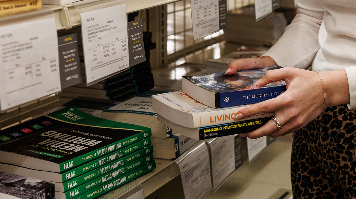 Close up photo of a woman's hands holding a stack of books in  front or a metal rack holding several stacks of books.