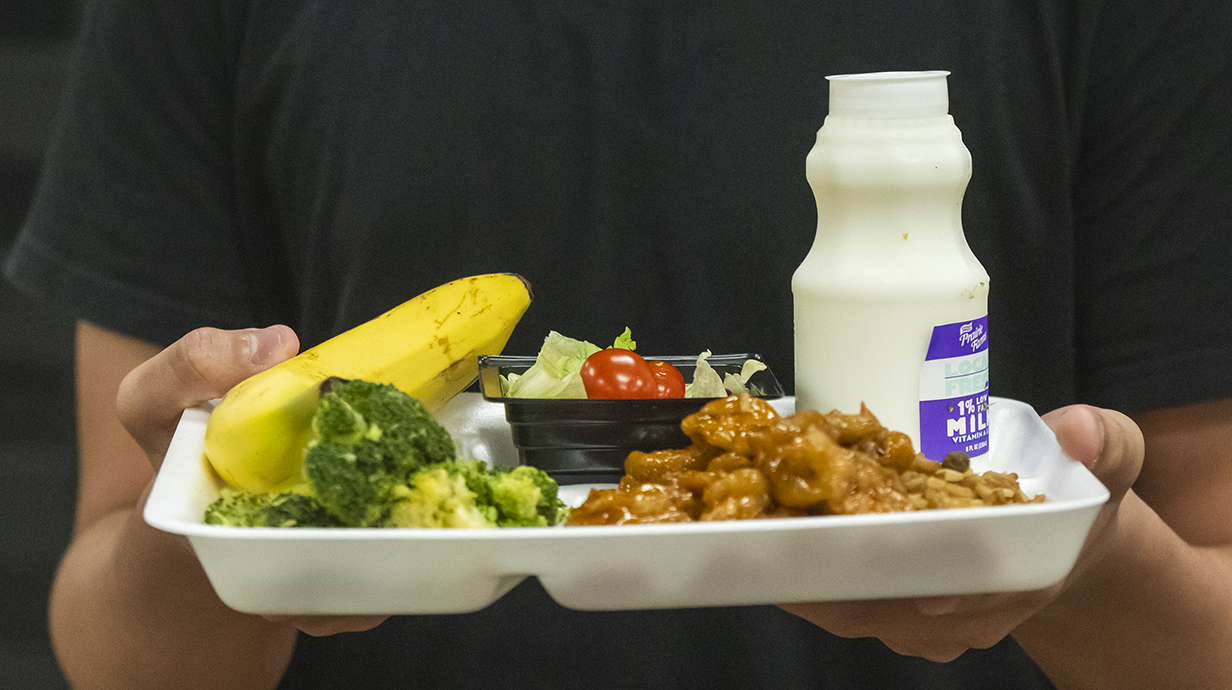 A student holds a lunch tray loaded with chicken, vegetables and a bottle of milk.
