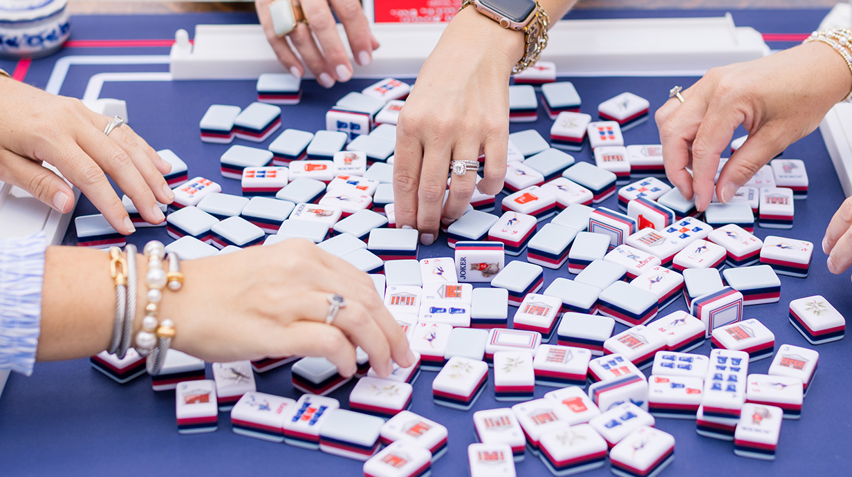 Several women's hands reach for mahjong tiles on a blue tabletop.
