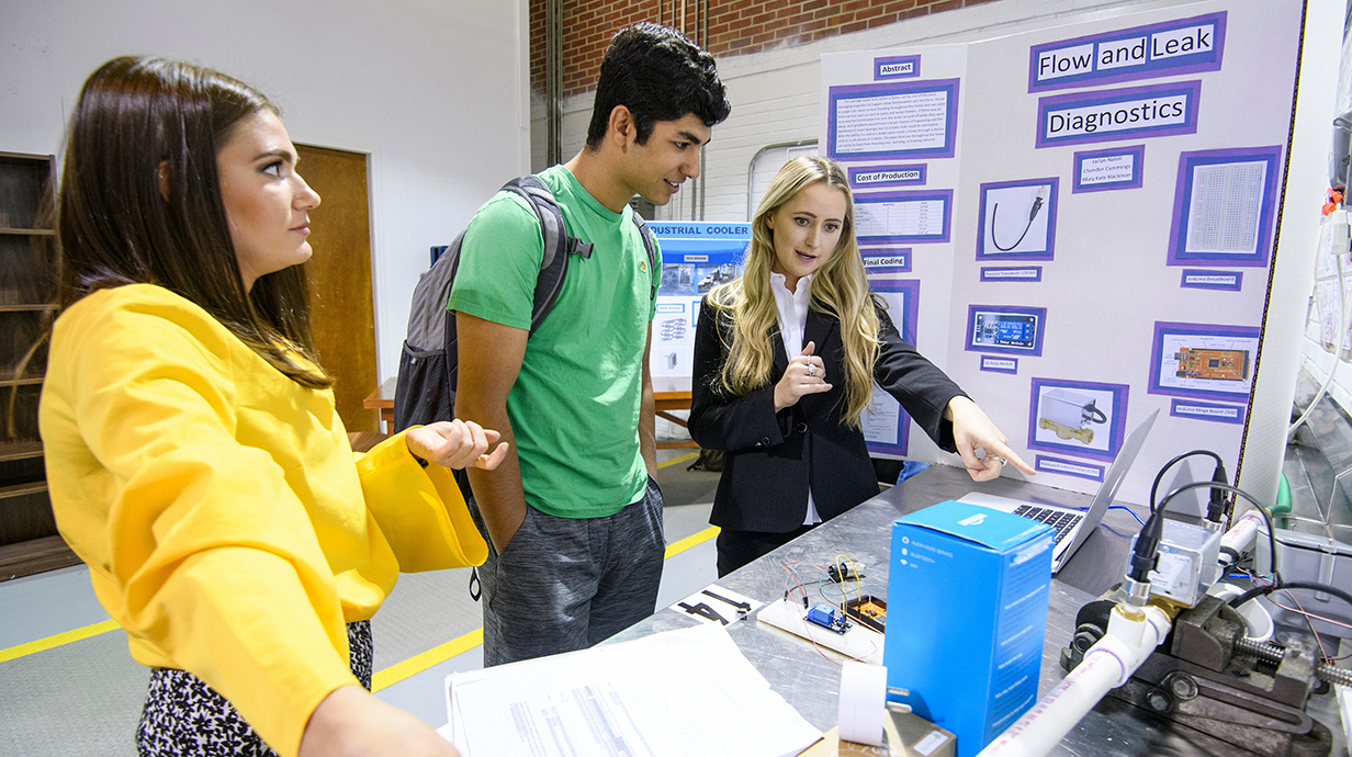 A young woman points to components in an engineering display while a young man and young woman watch.