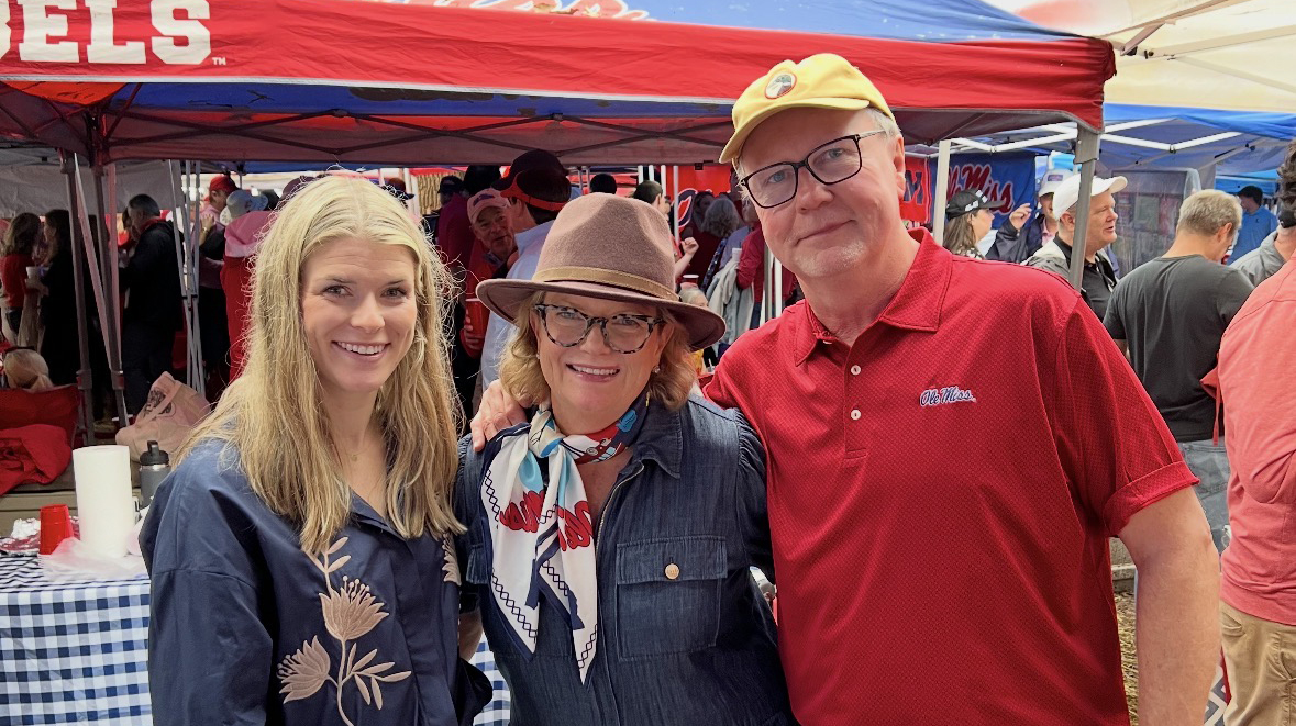 A man stand with two women, all wearing Ole Miss gear, underneath a pop-up tent in a tree-filled park.
