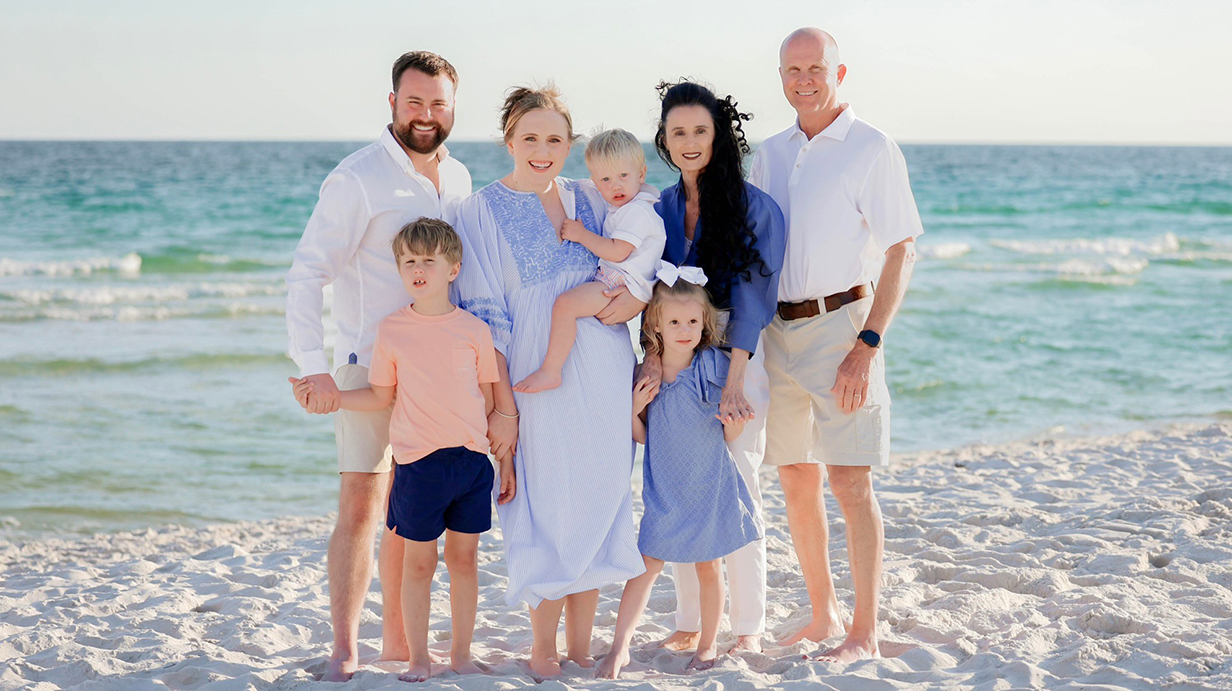 Two men, two women, two young boys and a young girl stand on a beach in front of the ocean.
