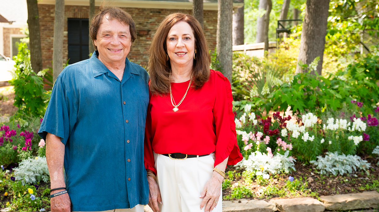 A man and woman stand outdoors in front of colorful flowers and shrubbery.