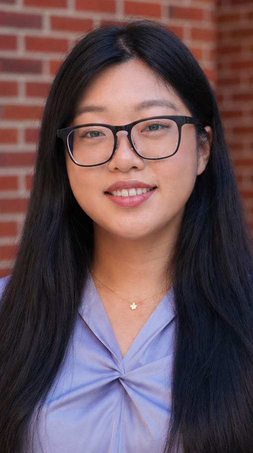 Headshot of a young woman wearing a blue blouse standing in front of a brick wall.