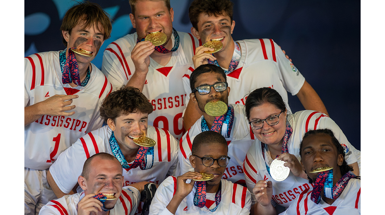 A group of young people, all wearing white-and-red football jerseys, bite down on large gold medals.