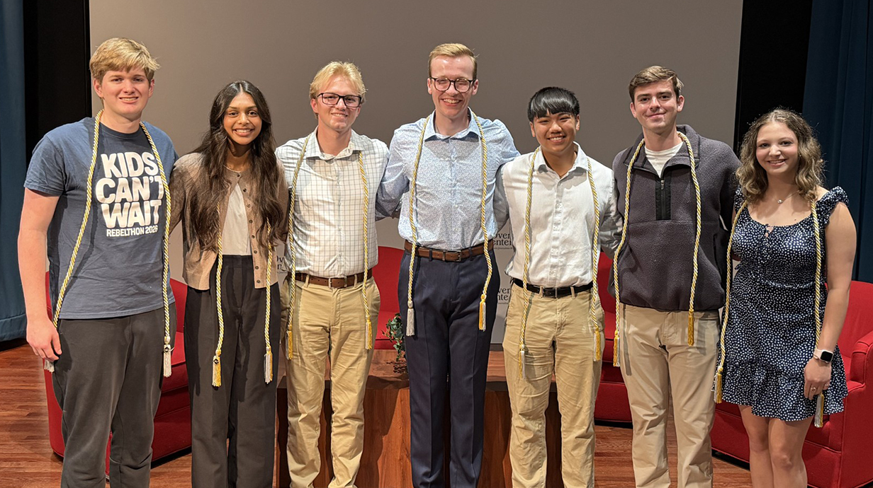 A group of young people, all wearing white graduation cords, stand on a stage.