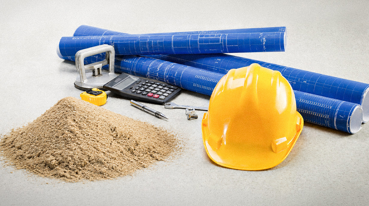 A yellow hardhat sits among a pile of rolled blueprints and a pile of sand.