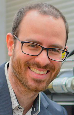 Headshot of a man wearing glasses and a blue-gray jacket.