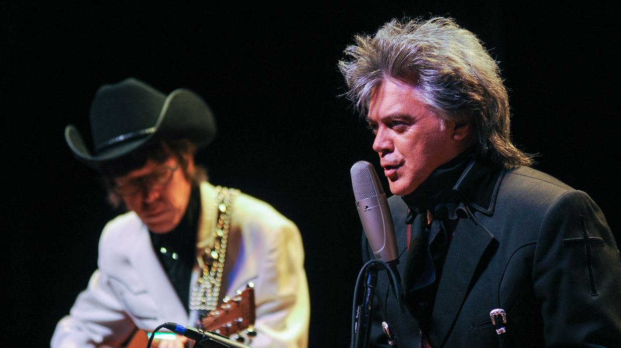 A man sings into a microphone while another man strums a guitar on a stage.
