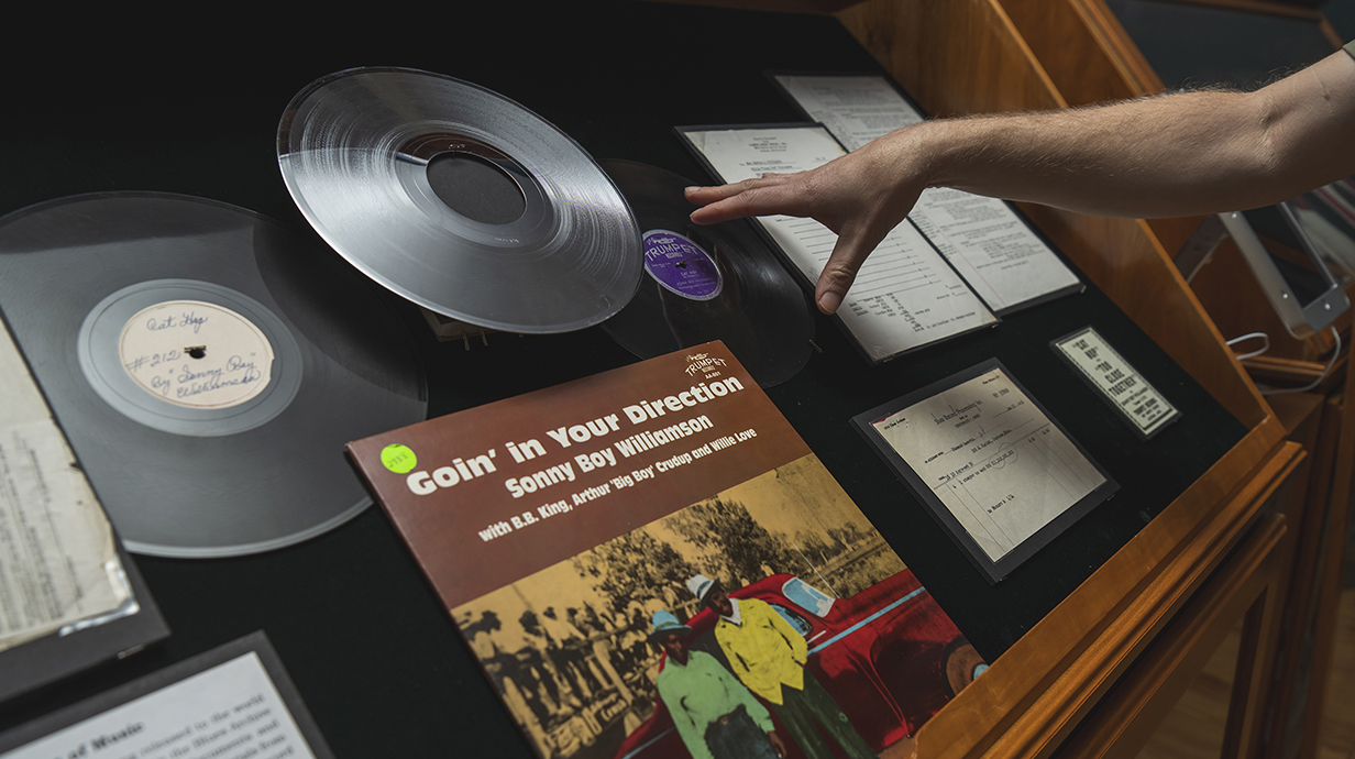 A hand points at a display case filled with old vinyl records, written correspondence and lyrics.