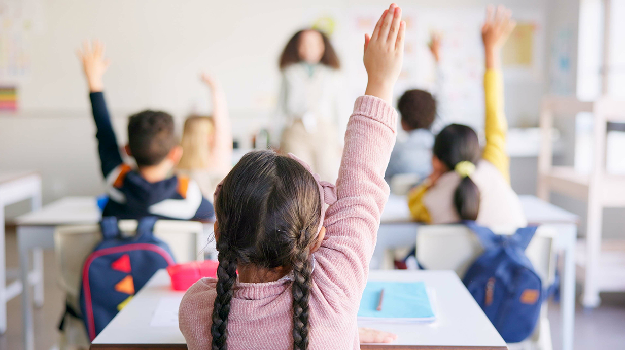 A young girl hold up her hand in a classroom full of students.