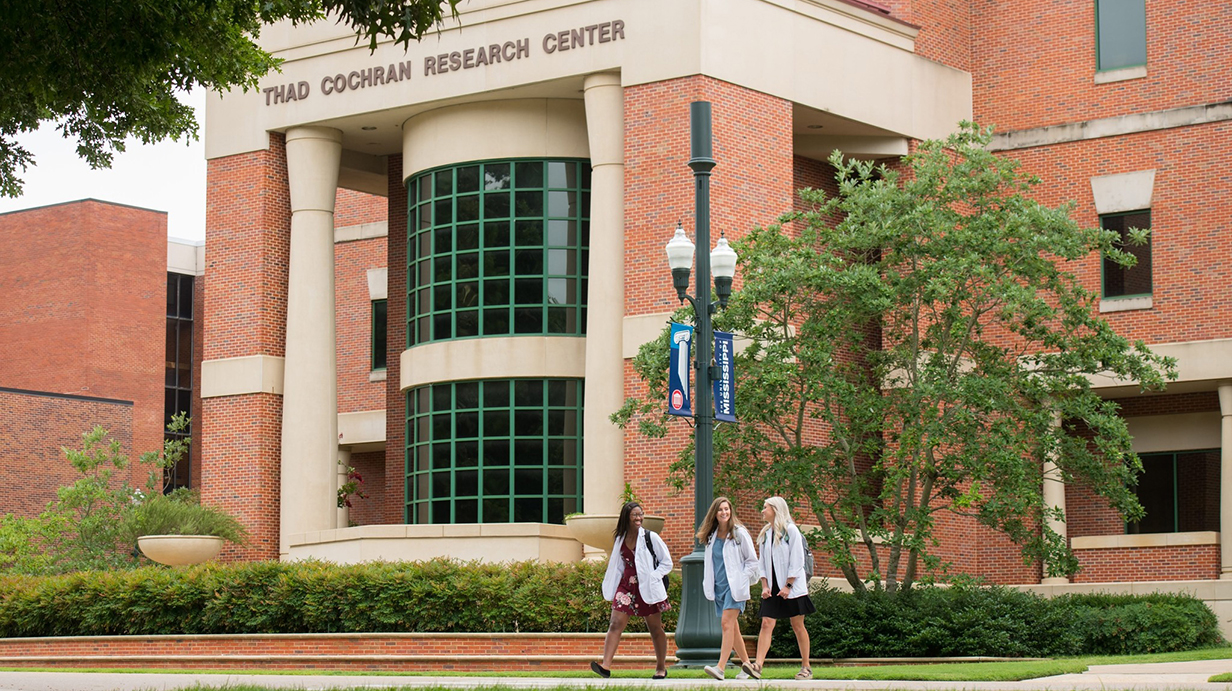 Three young women wearing white lab coats walk in front of a large brick building with 'Thad Cochran Research Center' along the top.