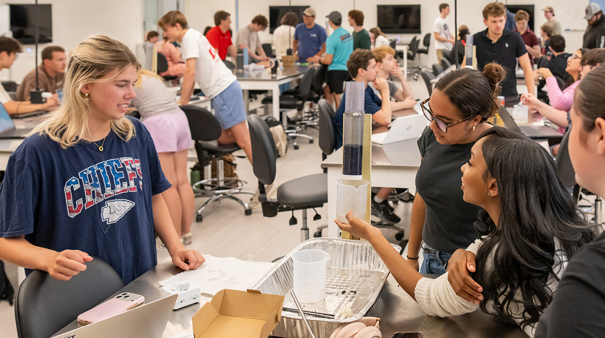 Three young women conduct an engineering experiment in a classroom filled with students clustered around lab tables.