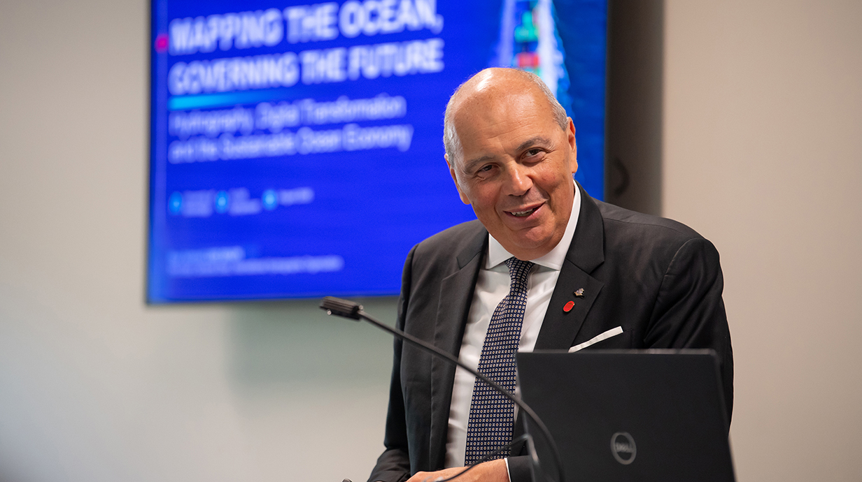 A man wearing a dark suit smiles while standing at a podium in front of a projection screen bearing a heading that reads 'Mapping the Ocean.'