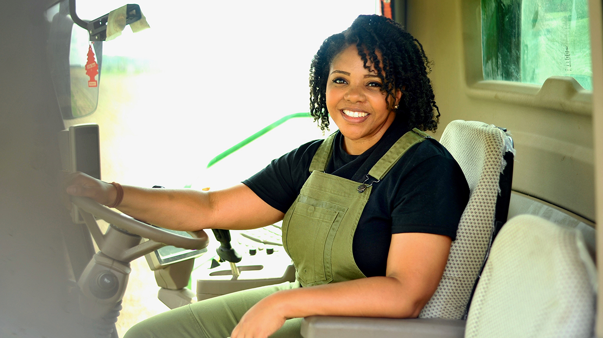 A woman wearing olive overalls sits in the enclosed cab of a piece of farm machinery.