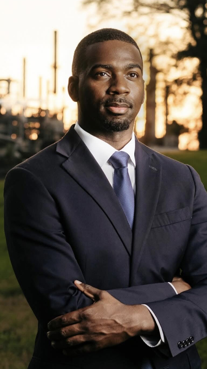 A young man wearing a suit stands in front of an industrial site.