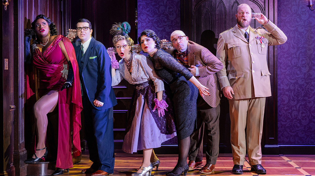 Three women and three men, all dressed in elaborate stage costumes, listen intently at a door in a wood-paneled hall.