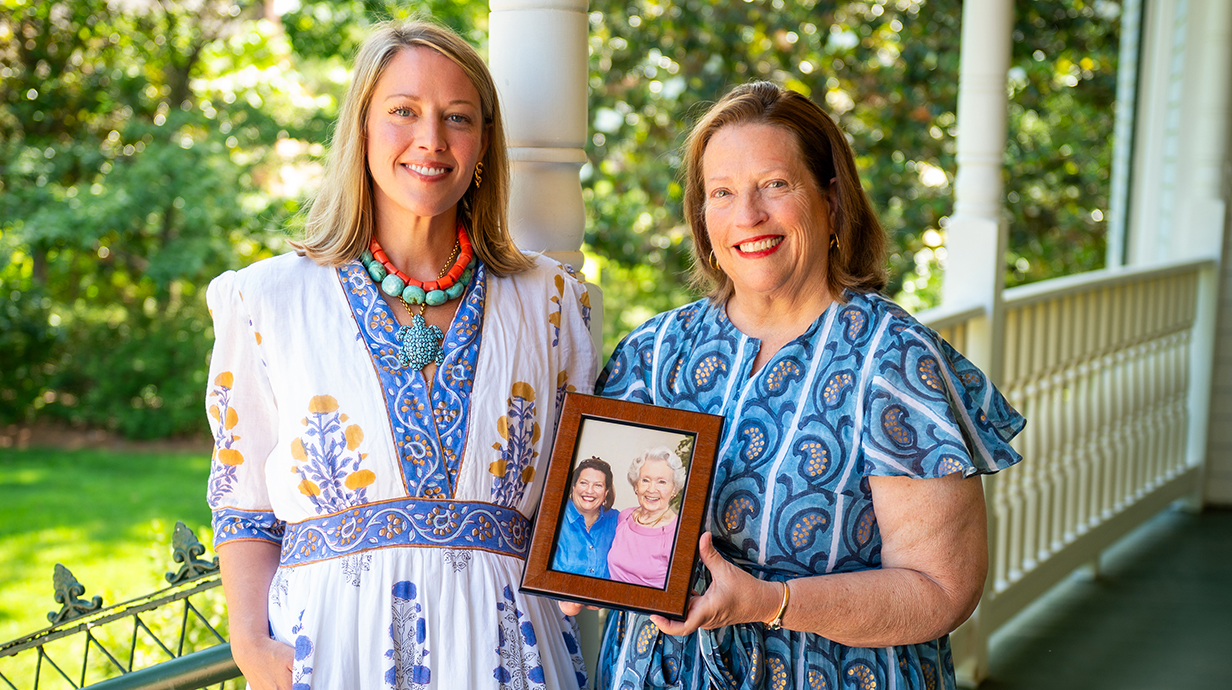 Two women stand on a covered porch holding a photograph of two women.
