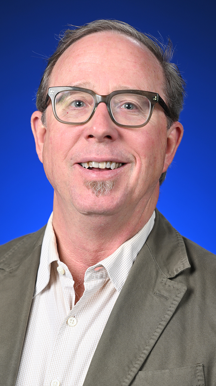 Headshot of a man wearing a tan jacket over an open-collared shirt.