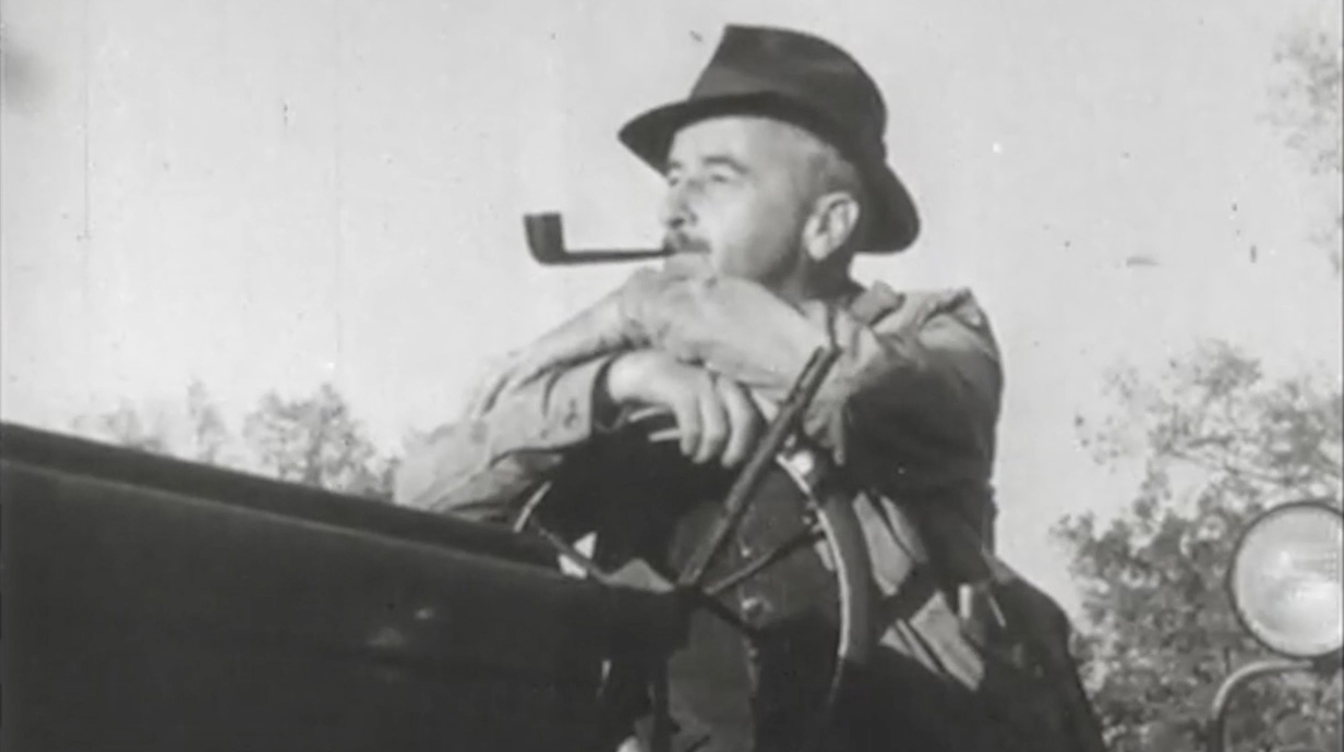 Black-and-white photo of a man wearing a hat and smoking a pipe while seated on a tractor.