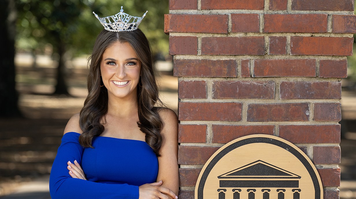A young woman wearing a blue dress and a tiara stands next to a brick column in a wooded park.