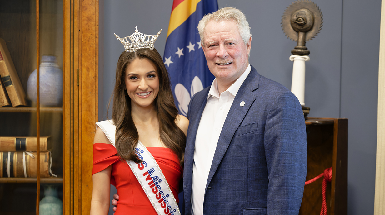 A young woman wearing a tiara and a sash reading 'Miss Mississippi" stands with a man wearing a blue jacket in an ornate office.