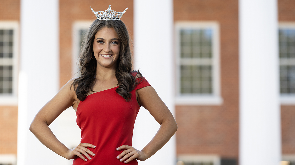 A young woman wearing a red dress and a tiara stands outside a large brick building with white columns.