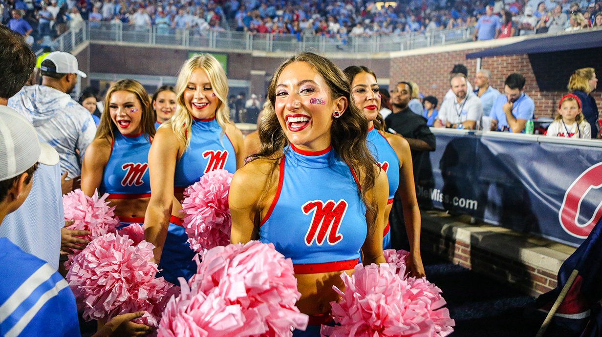 Several dancers wearing light blue uniforms with a red Ole Miss logo smile and cheer in a football stadium.