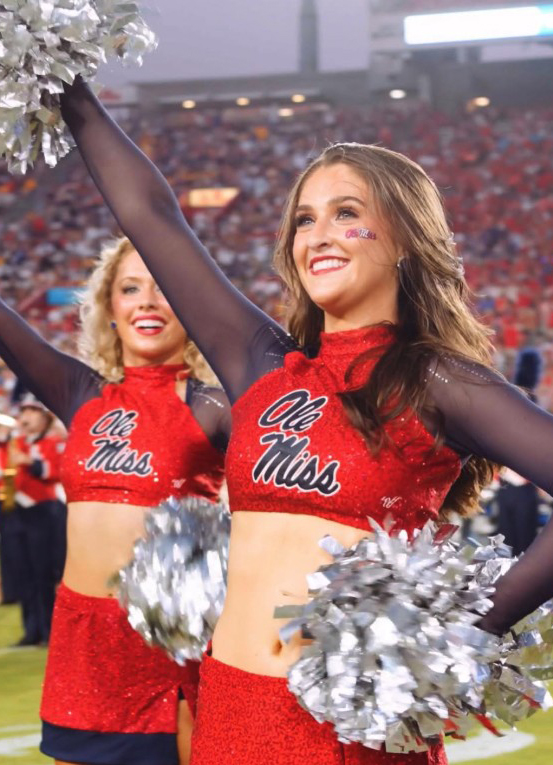 Two cheerleaders wave to teh crowd in a football stadium.