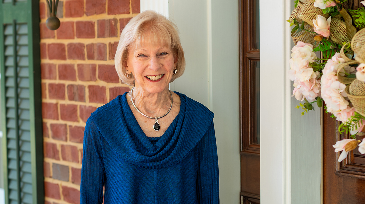 A woman wearing a blue sweater stands on the front porch of a brick house with a floral wreath on the door.